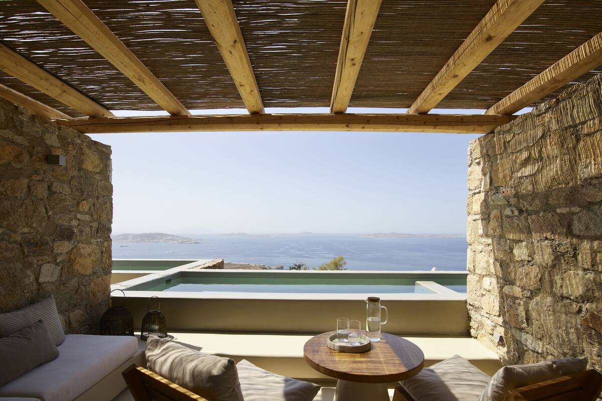 A room with a stone wall, a wooden ceiling, a view of the sea, a small pool, and a table with chairs.