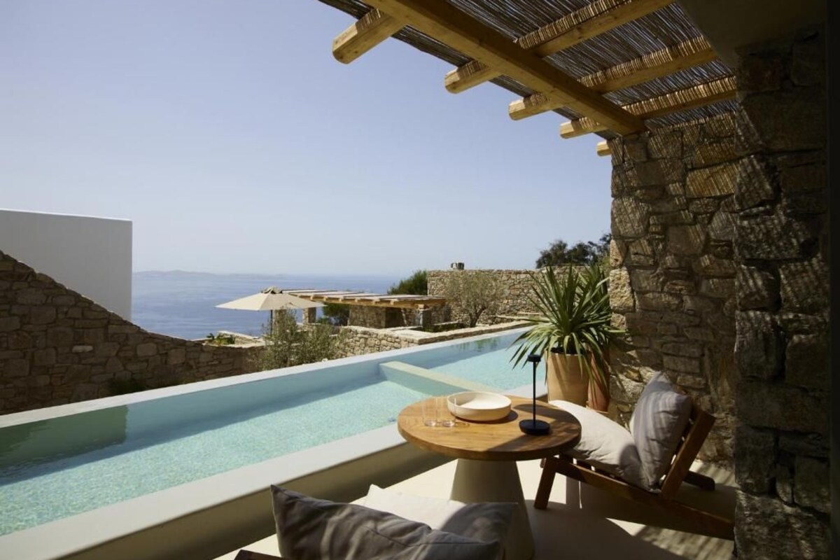 A private pool with a clear view of the sea, stone walls, a wooden pergola, and a round table with a bowl and a glass.