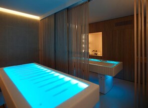 A modern spa room with a heated massage table, a wooden wall panel, and a white sink with a mirror.