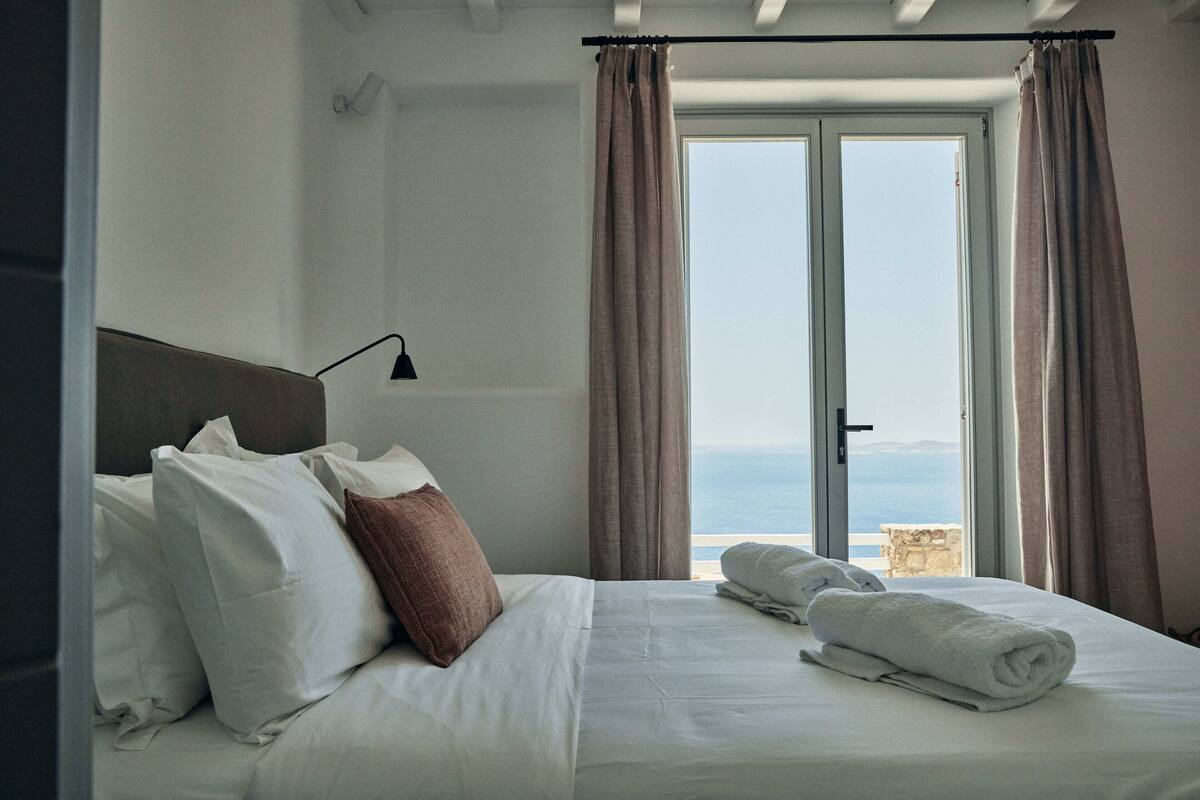 A neatly made bed with white and brown pillows, a folded towel, and a view of the sea through a window with curtains.