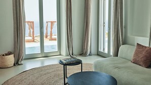 A room with a sofa, a small table with a book, and a view of an outdoor area with a pool and chairs. - Collini Hotel Mykonos, WorldHotels Elite (Mykonos)