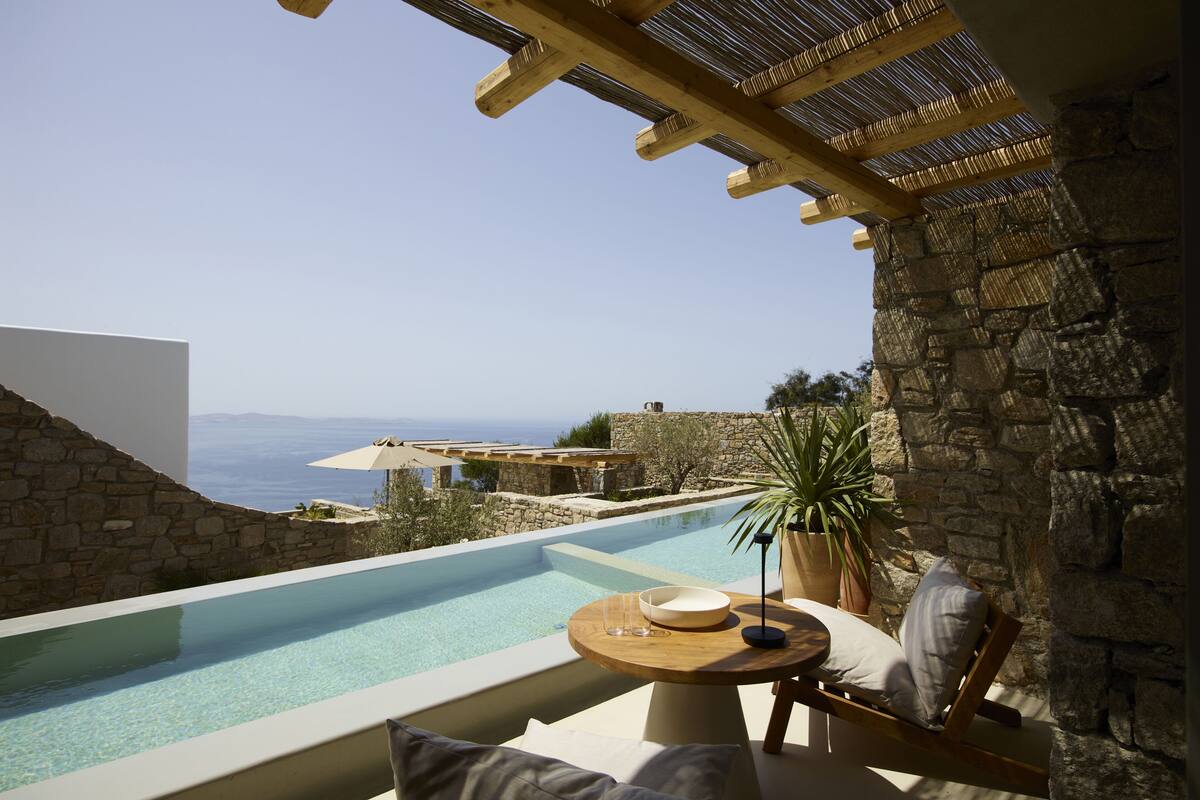 A stone-walled infinity pool with a clear view of the sea, a wooden pergola, and a round table with a bowl and glasses.