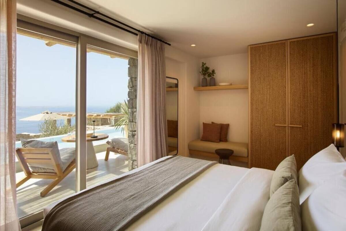 A bedroom with a large bed, a view of the sea, a balcony with a chair, and a wooden wardrobe.