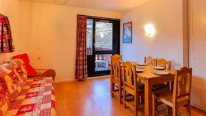 Ein gemütliches Zimmer mit Holztisch, Stühlen, einer gesteppten Couch, einem Fenster mit Vorhängen und einer Deckenleuchte.