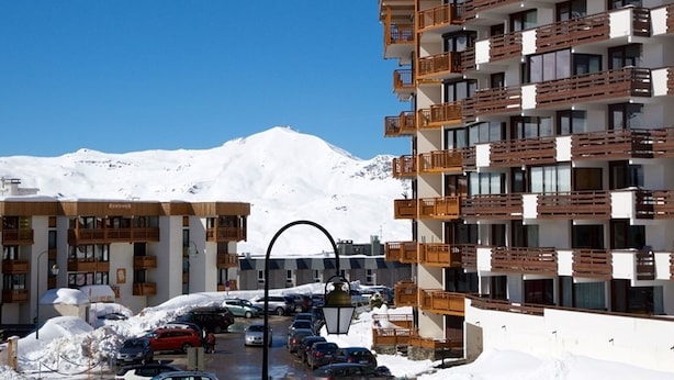Ein mehrstöckiges Hotel mit Balkonen, einem Parkplatz und einem verschneiten Berg im Hintergrund.