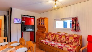 Ein gemütliches Zimmer mit einer Holzcouch, einem Esstisch mit Stühlen, einer Küchenzeile mit roten Schränken und einem Fenster mit Vorhängen.