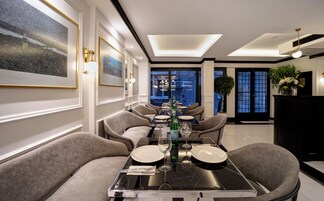 A dining area with a long table set for four, surrounded by upholstered chairs. There are framed artworks on the walls, a potted plant, and a black console table with a vase of flowers.