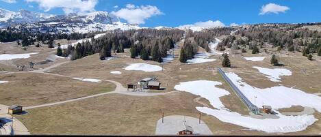 A mountainous landscape with snow-covered peaks, a clear sky, and a ski resort area with buildings and pathways.