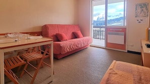 A cozy room with a dining table set for two, a red sofa, and a large window offering a mountain view. - Cozy Studio for 3 with Free WiFi, Close to Slopes & Shops, Private Ski Locker, Parking Available (Le Dévoluy)