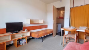 A compact hotel room with a single bed, a small desk with a chair, a television, a bookshelf, and a kitchenette. - Cozy Studio for 3 with Free WiFi, Close to Slopes & Shops, Private Ski Locker, Parking Available (Le Dévoluy)