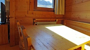A wooden cabin with a large table and benches, a window with a view of mountains, and a radiator under the table.