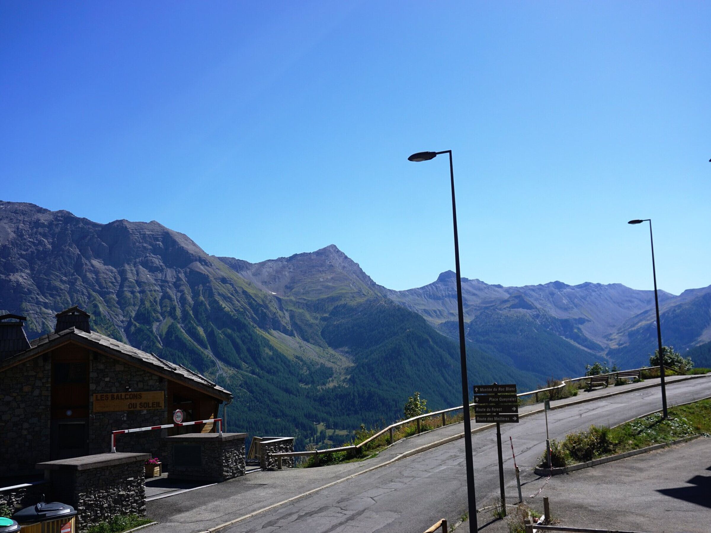 A mountain lodge with a stone facade, a sign indicating 'Les Balcons Ou Soleil', and a road leading to mountain trails.