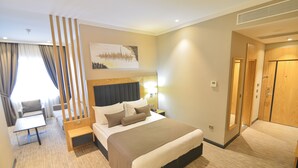 A hotel room with a large bed, bedside tables, a desk, and a chair. There is a window with curtains, a painting on the wall, and a door to the right. - Serenity Comfort Hotel  (Istanbul)