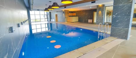 An indoor swimming pool with a blue tiled floor, a reception desk, and modern pendant lighting.