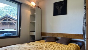 Un dormitorio con una cama, un ventanal con vistas a una cabaña, una obra de arte en la pared que representa una cabeza de ciervo y un cabecero de madera.