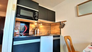 Cocina compacta con horno empotrado, lavadero y un refrigerador pequeño. Hay una silla de madera y una mesa con mantel.