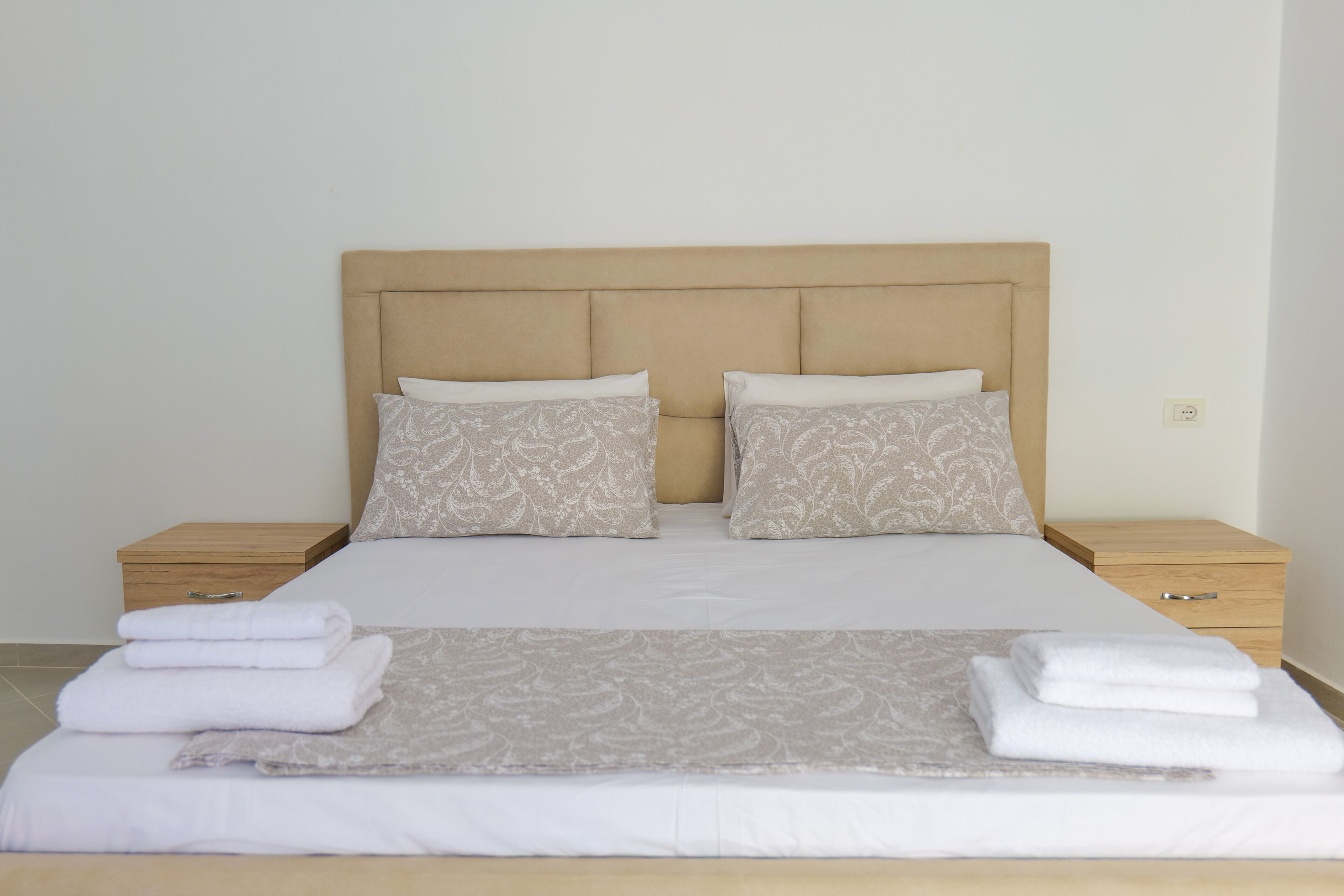 Una cama bien hecha con un cabecero color crema, dos almohadas iguales y una toalla blanca doblada sobre la cama.