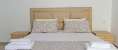 Una cama bien hecha con un cabecero color crema, dos almohadas iguales y una toalla blanca doblada sobre la cama.