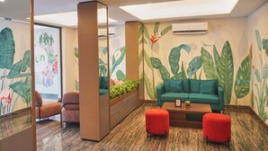 A modern room with a green sofa, red ottomans, a wooden cabinet with plants, and a mural of tropical leaves. - Lun Hotel Manado (Manado)