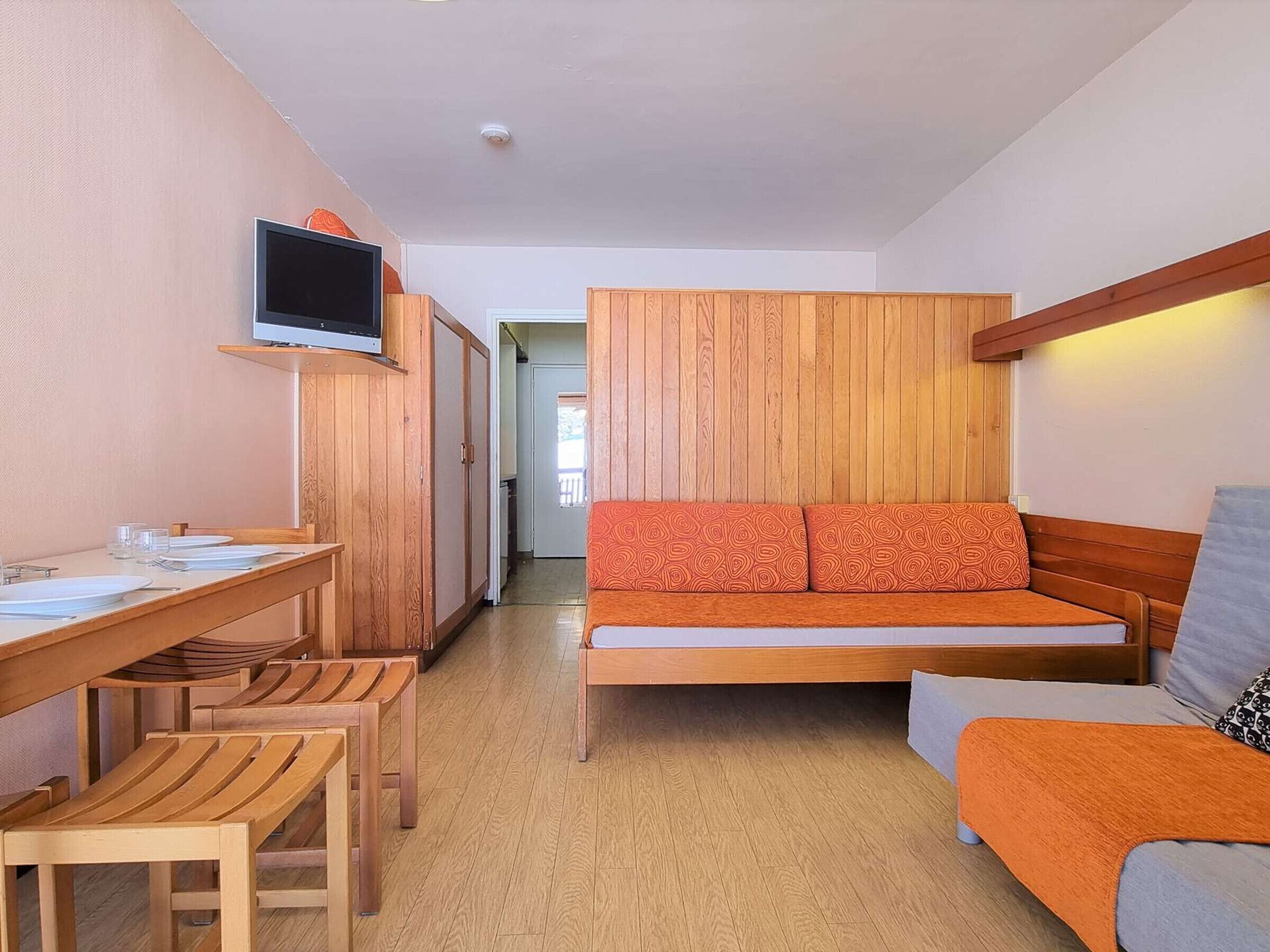 Une pièce de taille modeste, avec un lit en bois, un téléviseur fixé au mur, une table à manger avec des chaises et un petit coin cuisine.