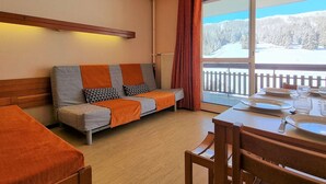 A cozy room with a sofa, a dining table set for two, and a large window offering a snowy landscape view. - Cozy studio with WIFI 50m from slopes - 4 ppl (Le Dévoluy)