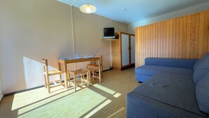Ein Zimmer mit Holzboden, einem blauen Sofa, einem Esstisch mit Stühlen, einem an einer Holzkommode befestigten Fernseher und einer Deckenleuchte.