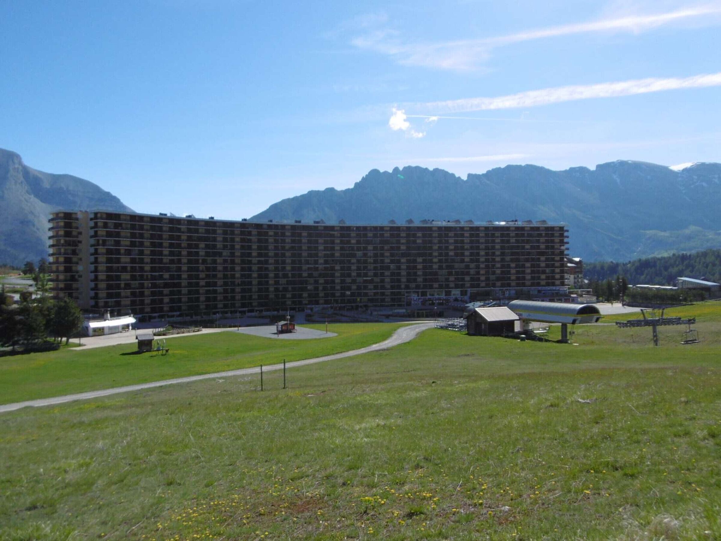 A large, multi-story hotel with a curved facade, situated in a grassy area with mountains in the background.