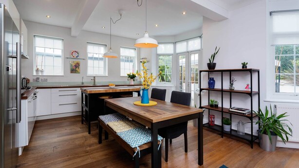 Une cuisine moderne avec une table à manger en bois, des bancs et des luminaires suspendus. On y trouve également une bibliothèque agrémentée de plantes et d’objets décoratifs.