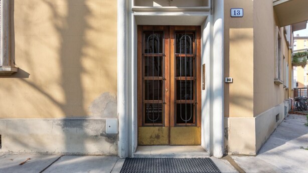 Une entrée d’immeuble avec une porte double en bois, une plaque portant le numéro « 13 » et un paillasson noir devant l’entrée.