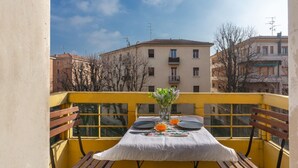 Un balcon avec une table dress ée pour deux personnes, un vase rempli de fleurs et une vue sur les bâtiments en arrière-plan.