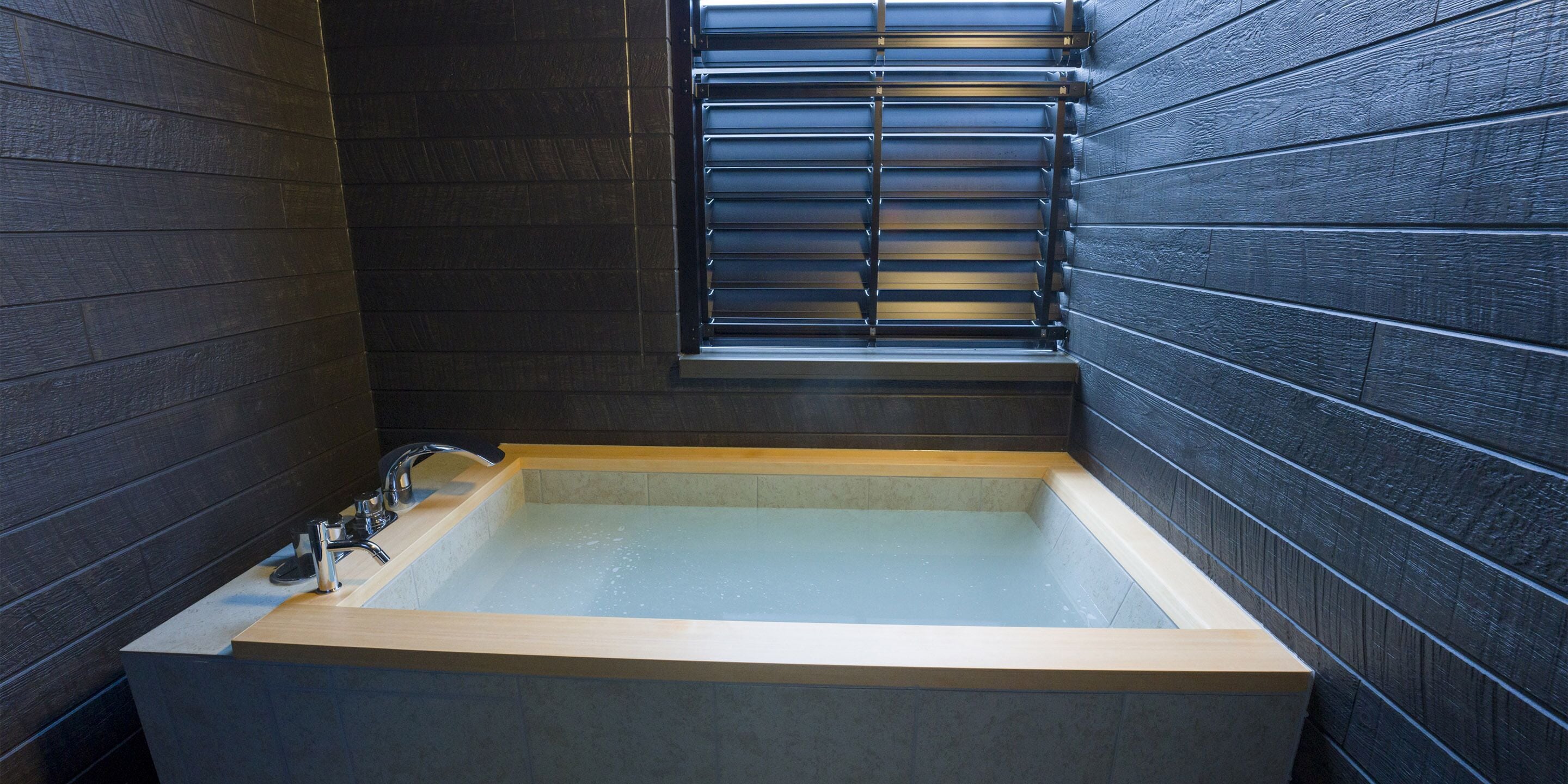 A modern bathroom with a freestanding bathtub, a window with blinds, and dark wooden walls.