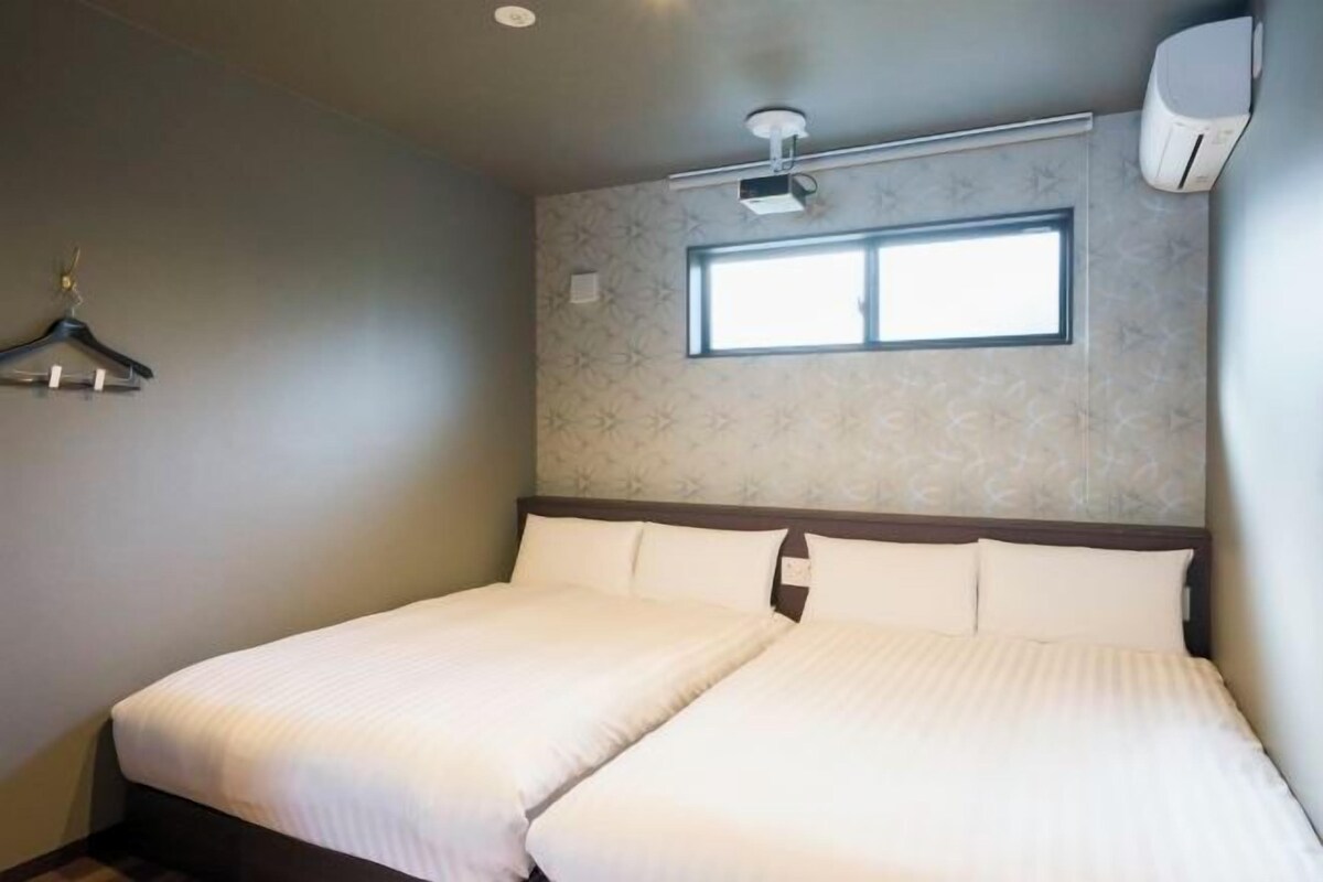 A double bed with white bedding, a window with blinds, a wall-mounted air conditioner, and a hanging coat rack.