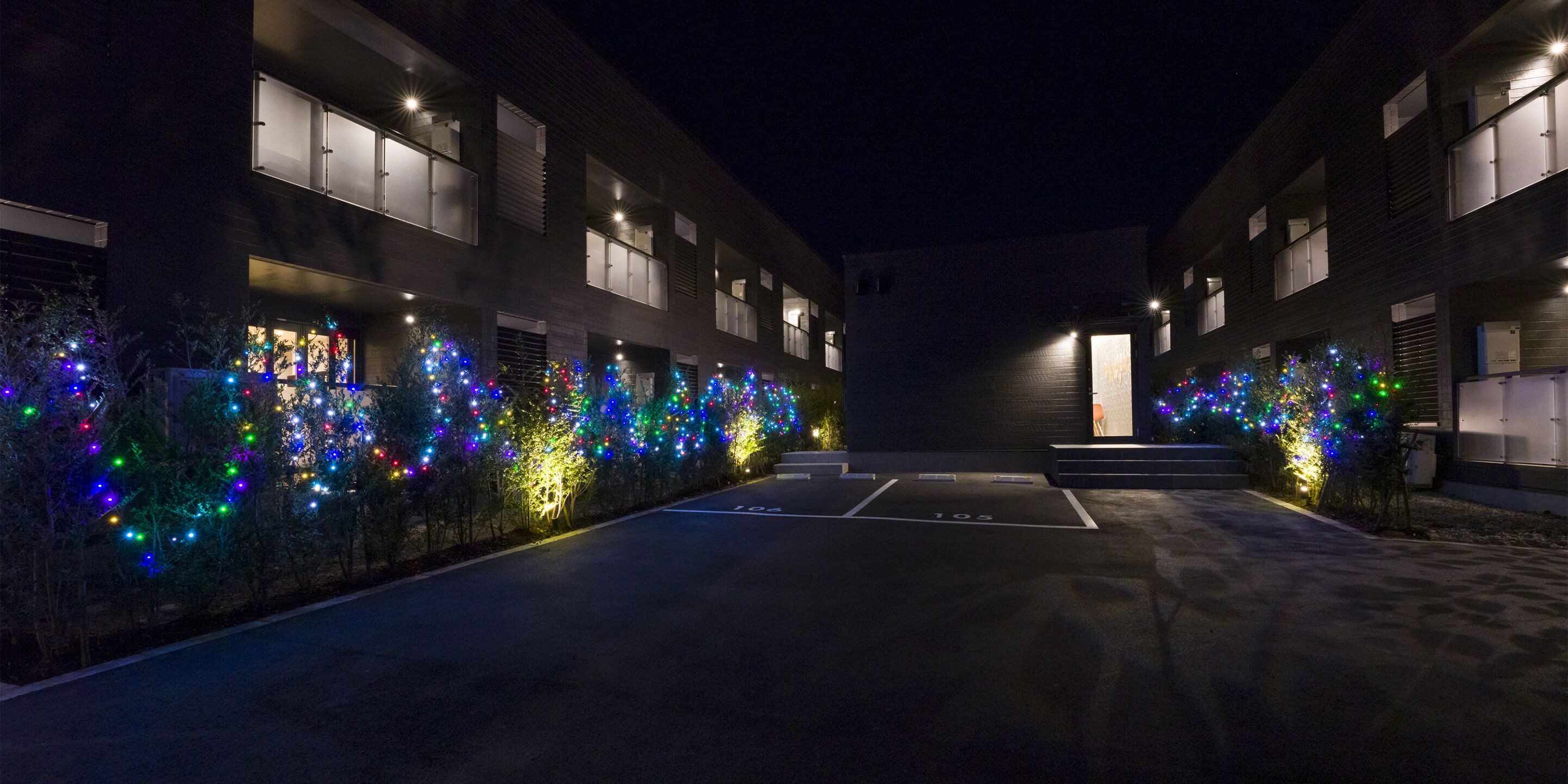 A parking area with trees decorated with colorful lights, a building with multiple windows, and a clear path marked with parking spaces.
