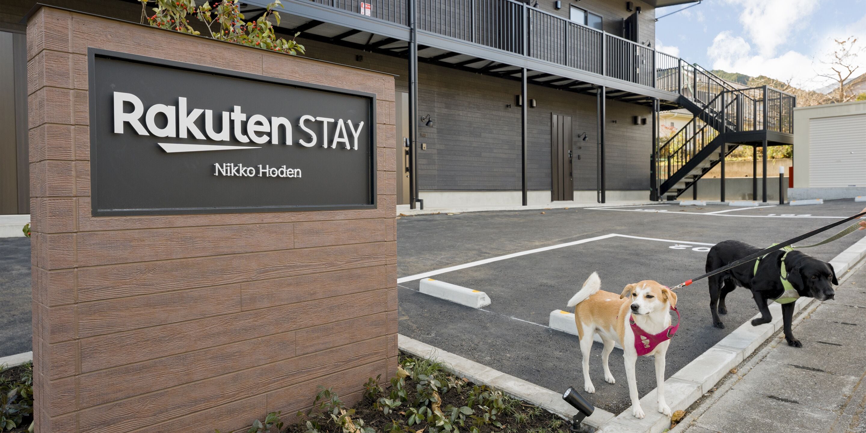 A sign for Rakuten Stay, a modern building with a wooden facade and a parking area.