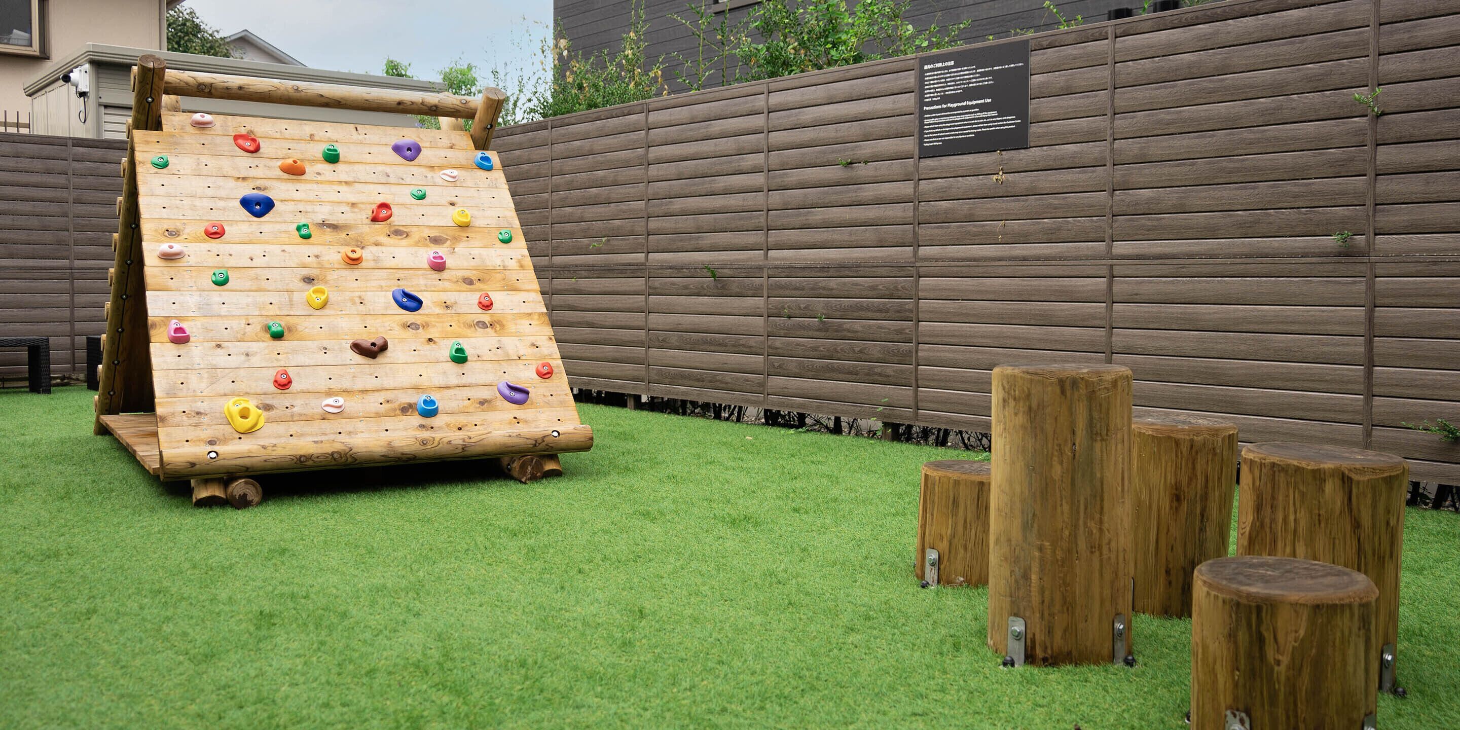 A wooden climbing wall with colorful holds, set on artificial grass, with wooden stumps and a wooden fence in the background.
