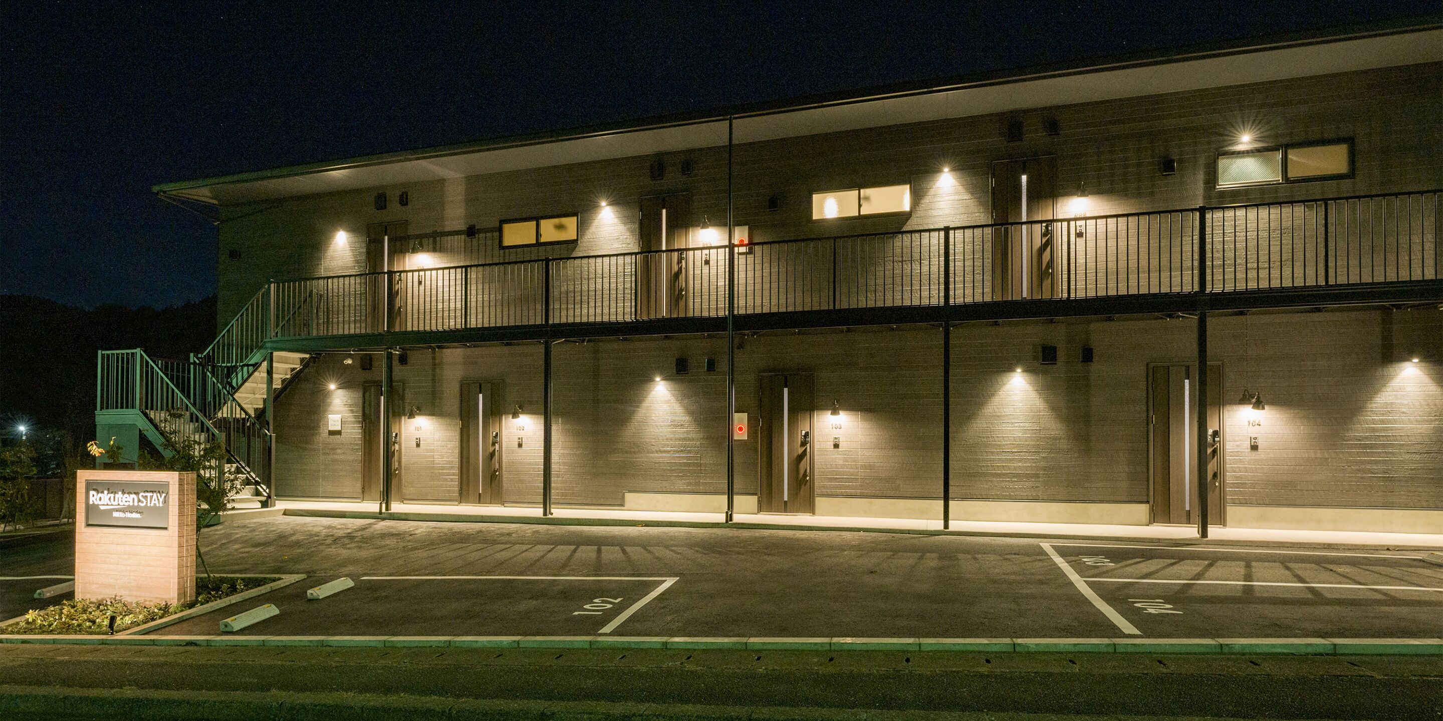 A two-story building with a parking lot, multiple doors, and a sign indicating it is a "Rakuten Stay" hotel.