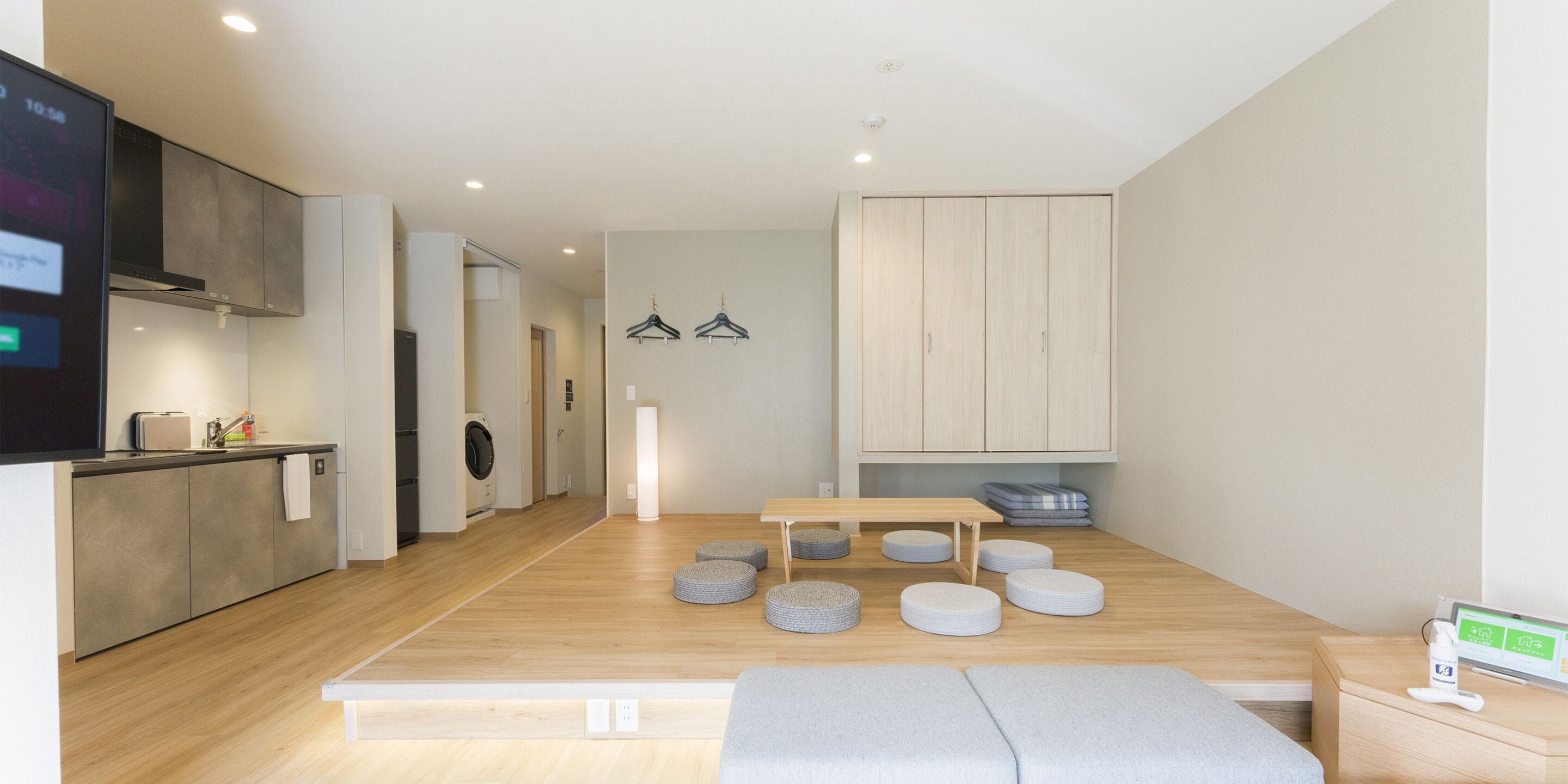 A modern, well-lit room with a wooden floor, a dining table, and a bench. There is a kitchen area with cabinets and a washing machine.