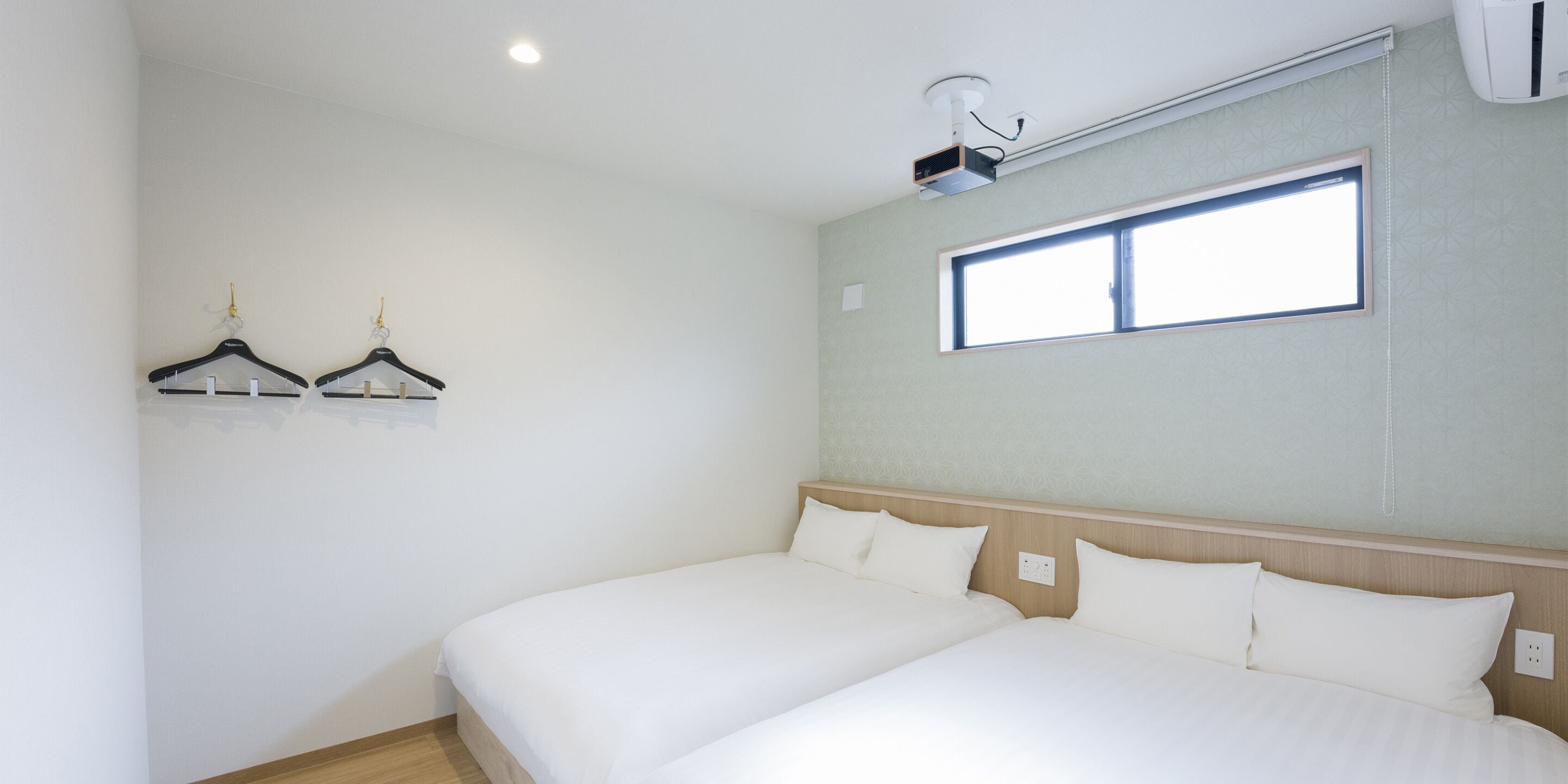 A hotel room with two beds, a ceiling projector, a window, and a wall-mounted air conditioner.
