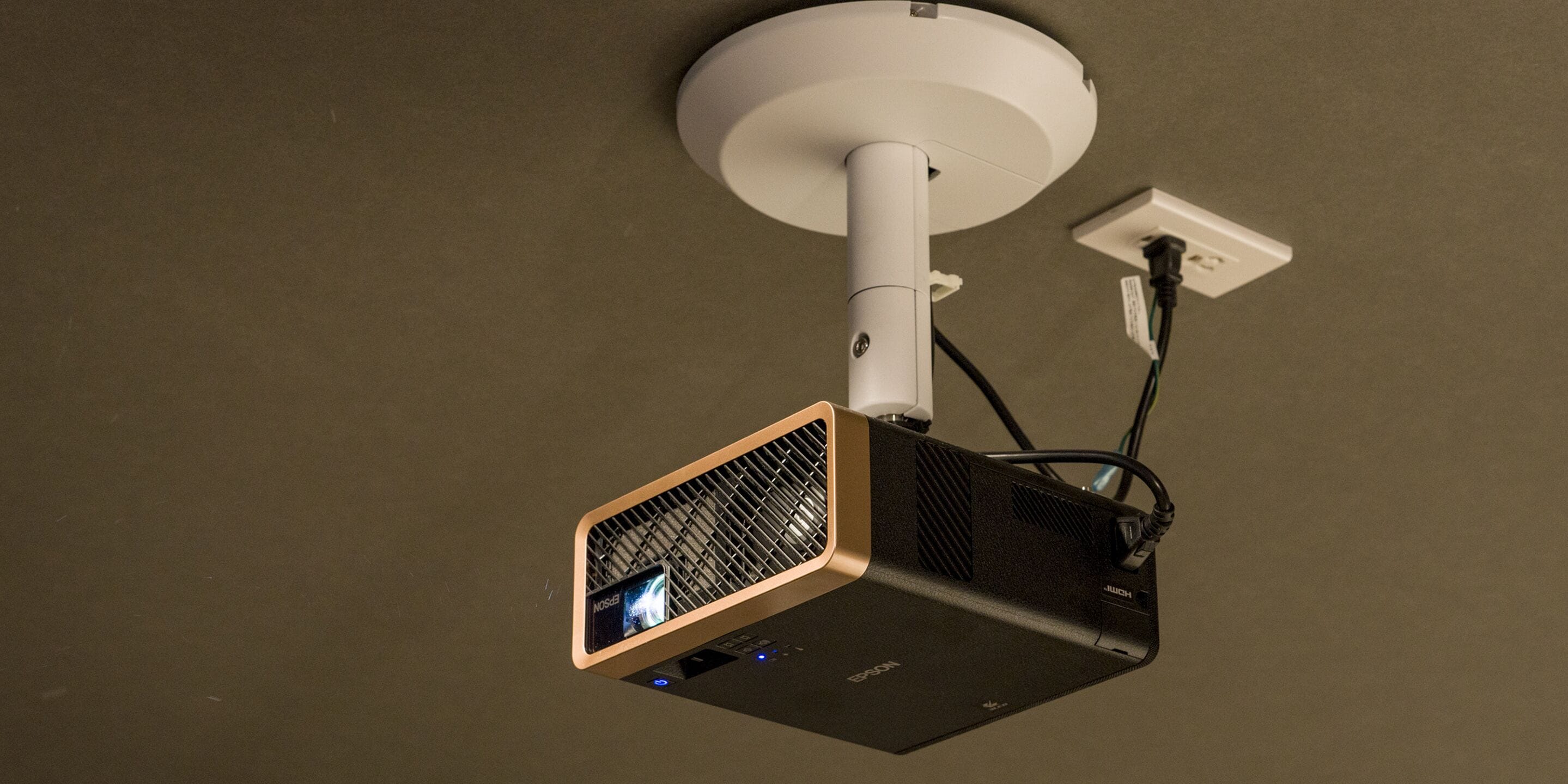 A ceiling-mounted projector with a white ceiling mount and a power cord plugged into a wall socket.