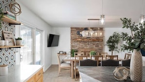 A modern kitchen with wooden cabinets, a dining area with a brick accent wall, and a TV mounted on the wall.