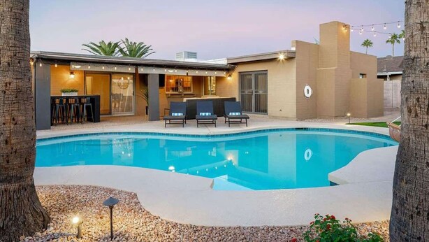 A modern outdoor pool area with a bar and seating, surrounded by a landscaped garden and a building with a flat roof.