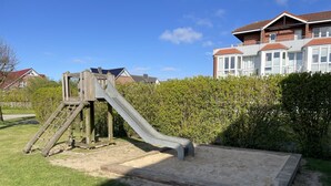 Ein Spielplatz mit Rutsche und Klettergerüst, umgeben von einer Rasenfläche und einem Heckenstreifen.