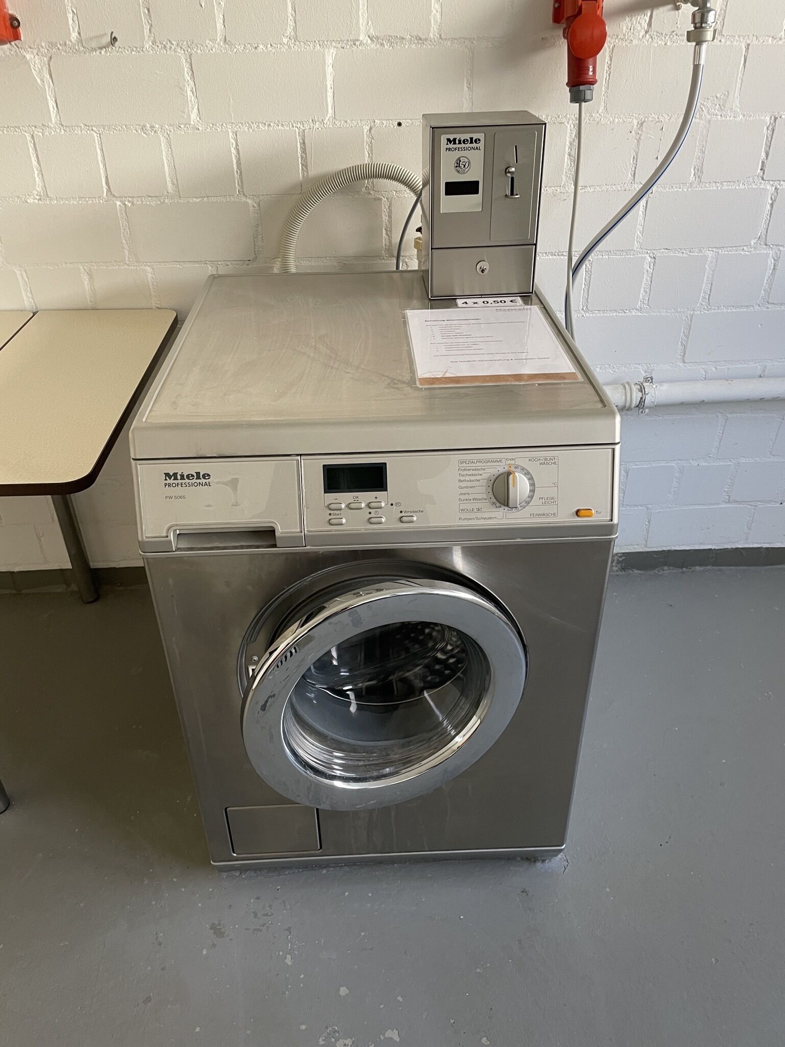 Eine kompakte, professionelle Miele Waschmaschine in einem Hauswirtschaftsraum mit weißer Ziegelwand und einem Tisch zur Linken.