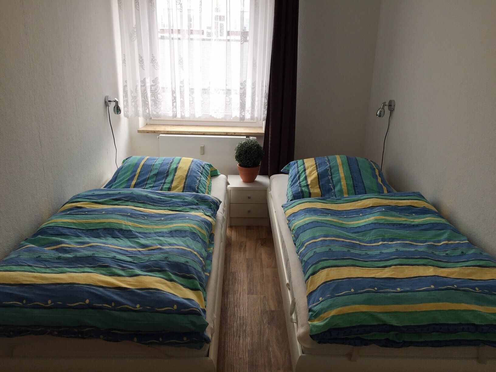 Una habitación pequeña y sencilla con dos camas, una ventana con cortinas traslúcidas, una pequeña mesa con planta en maceta y un aplique de pared.