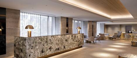 A modern hotel lobby with a marble reception desk, seating area, and large windows.