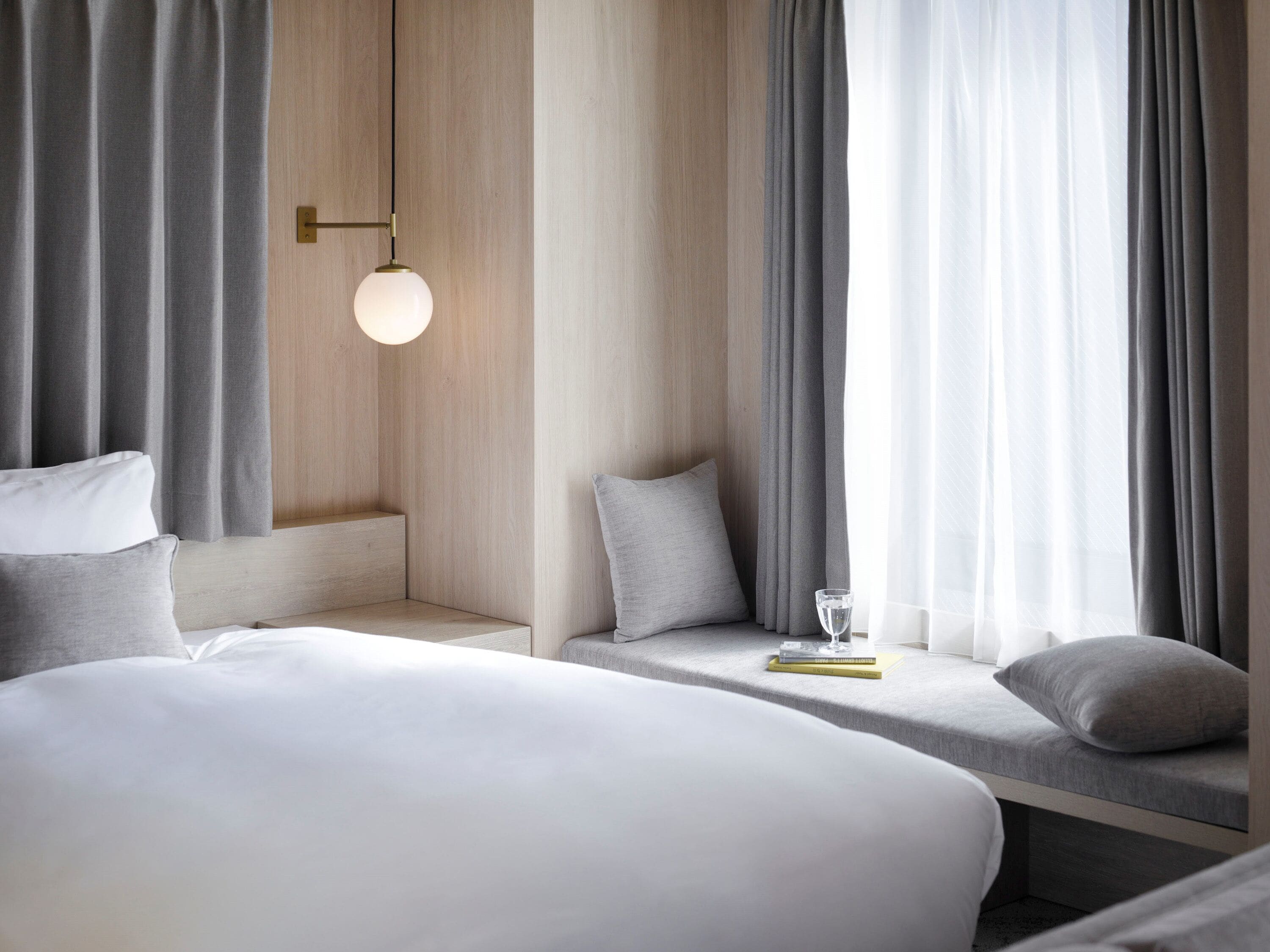 A modern hotel room with a large bed, a bedside table, a reading area with a chair, and a large window with curtains.