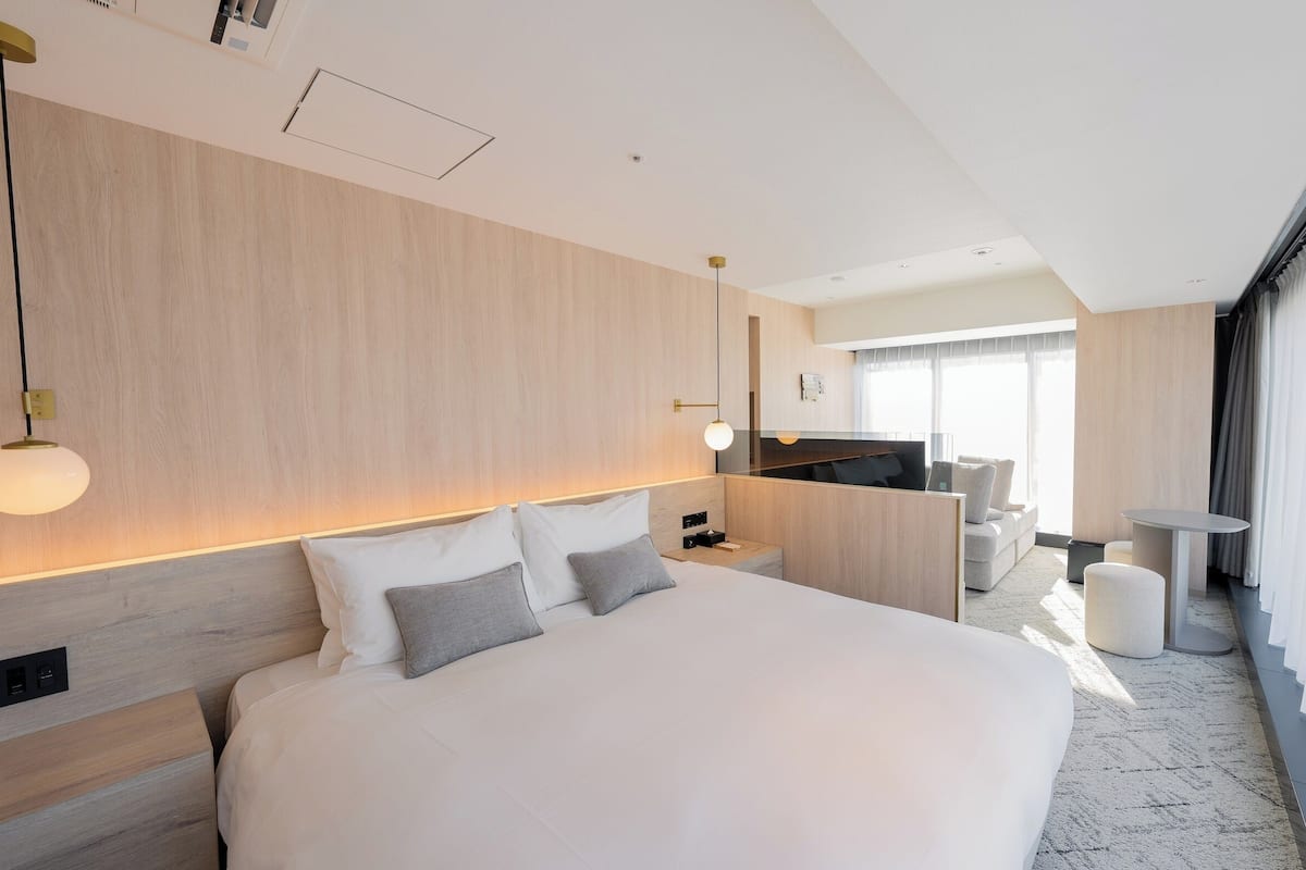 Quarto de hotel moderno com uma cama grande, mesas de cabeceira, escrivaninha, cadeira e vista para a cozinha e a sala de estar.