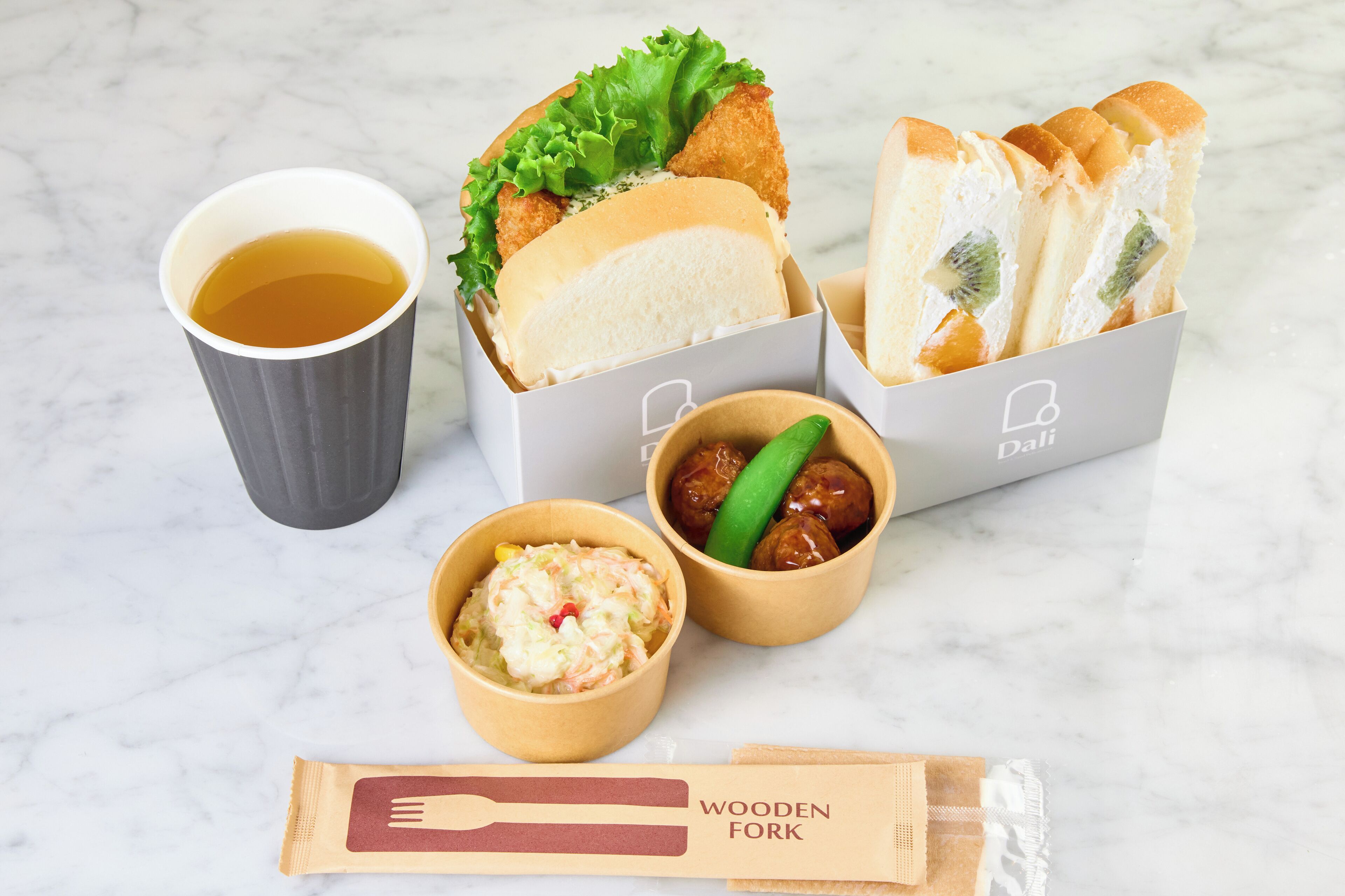 Une table avec une tasse de thé, du pain, un petit bol de salade de chou et un paquet de fourchettes en bois.