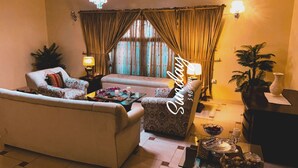 Ein Wohnzimmer mit Sofa, Sesseln und einem Couchtisch mit Dekoration. Eine Lampe steht auf einem Beistelltisch, und im Raum steht eine kleine Pflanze.