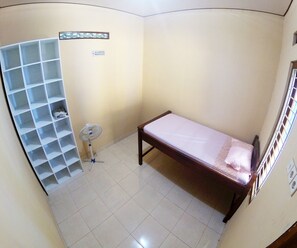 A small, single-bed room with a wooden bed frame and a white mattress. There is a white shelving unit with a few items on the lower shelves. A standing fan is placed near the shelving unit. The room has plain walls and a tiled floor.